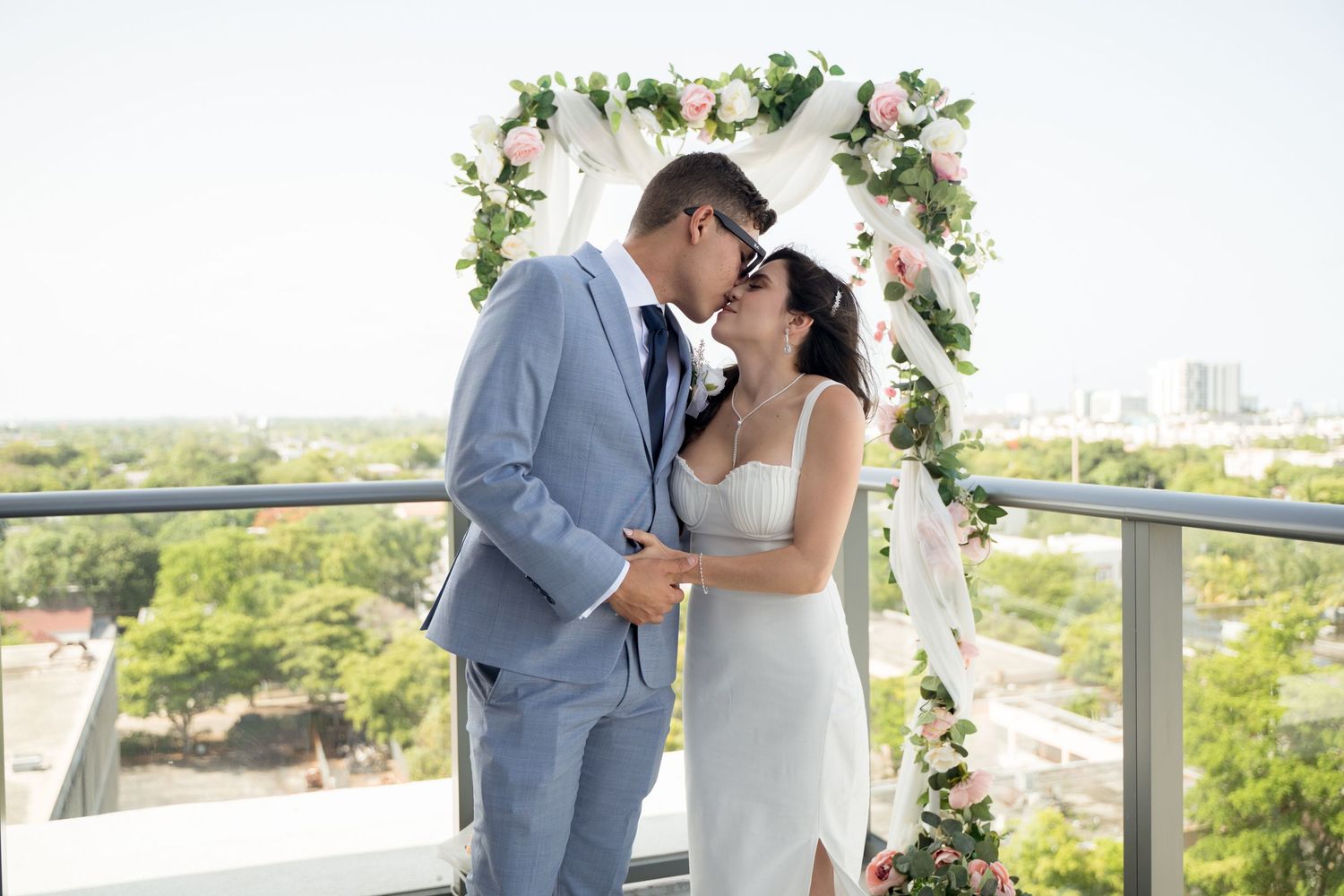 A romantic wedding couple shares kisses under a floral archway in an outdoor rooftop venue overlooking scenic greenery.
