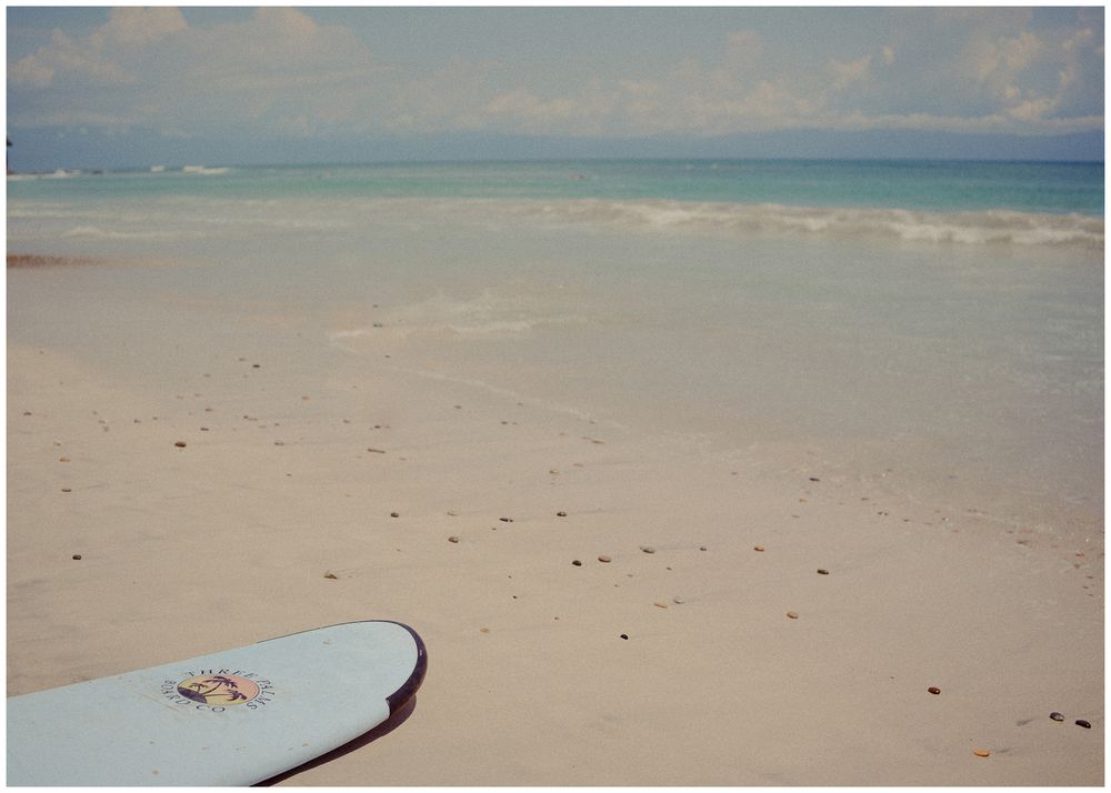 Wide sandy beach stretching to the horizon with calm turquoise waters and scattered rocks on the shore.