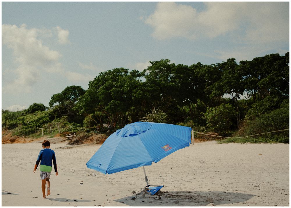 A person walks past a bright blue camping tent set up on a sandy beach with trees in the background.