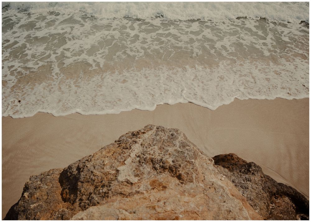 Rocky outcrop overlooking gentle waves washing up on a sandy beach shore.