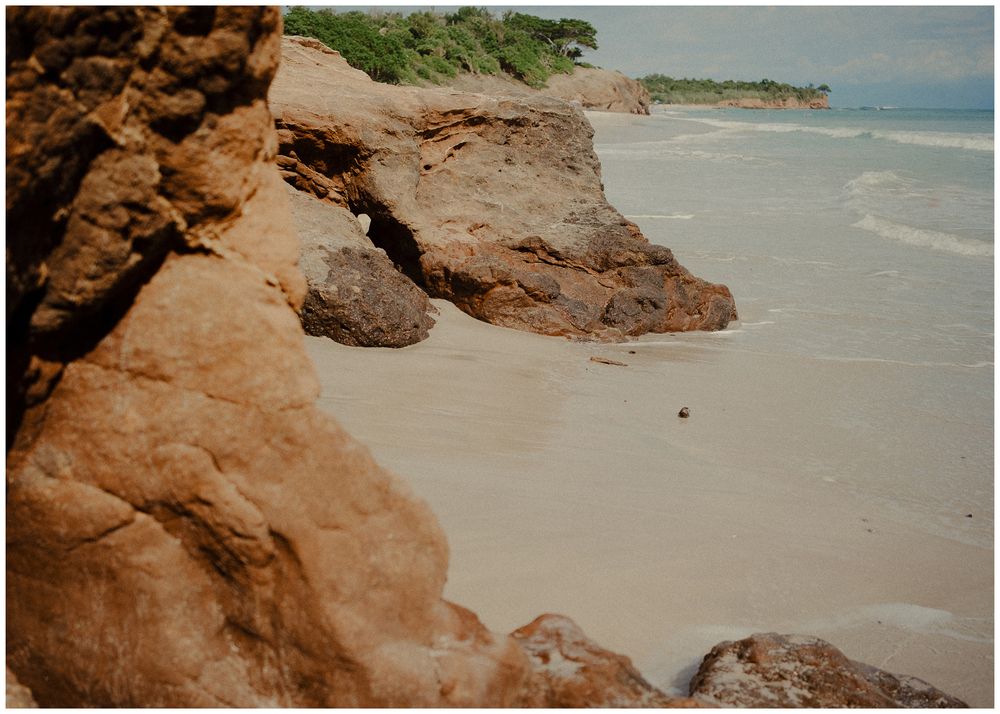 Scenic coastal landscape showing beige rocky cliffs meeting a white sandy beach with calm ocean waters.