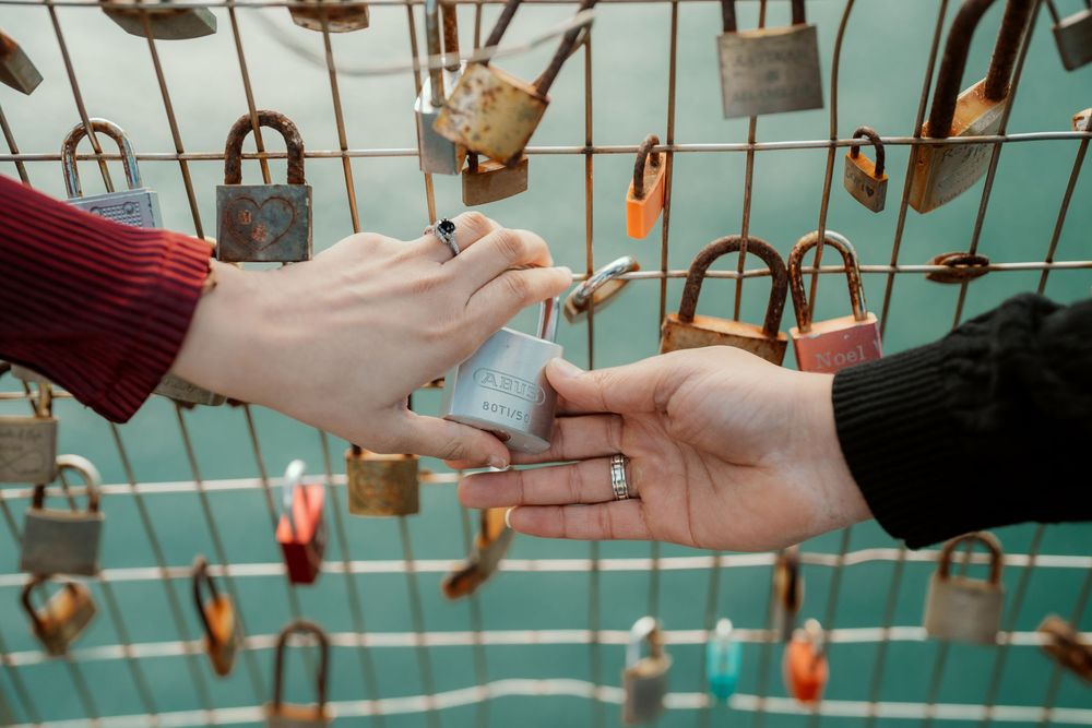 Two hands exchanging a love lock with numerous padlocks hanging in the background on turquoise-colored bars.