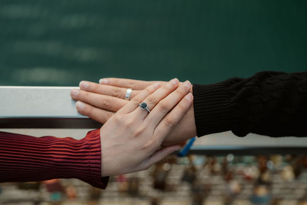 Two hands wearing rings touch on a railing against a dark green blurred background.