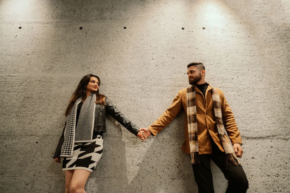 A couple in stylish fall outfits holds hands while standing against a concrete wall.