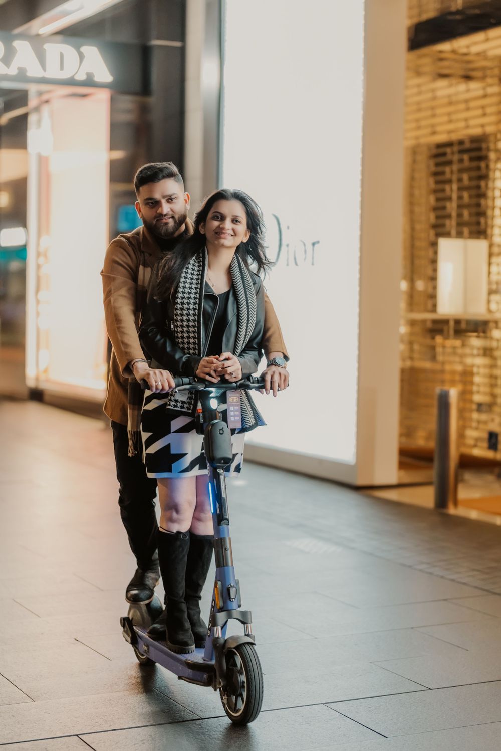 Two people ride an electric scooter together through a modern shopping mall with Prada store in background.