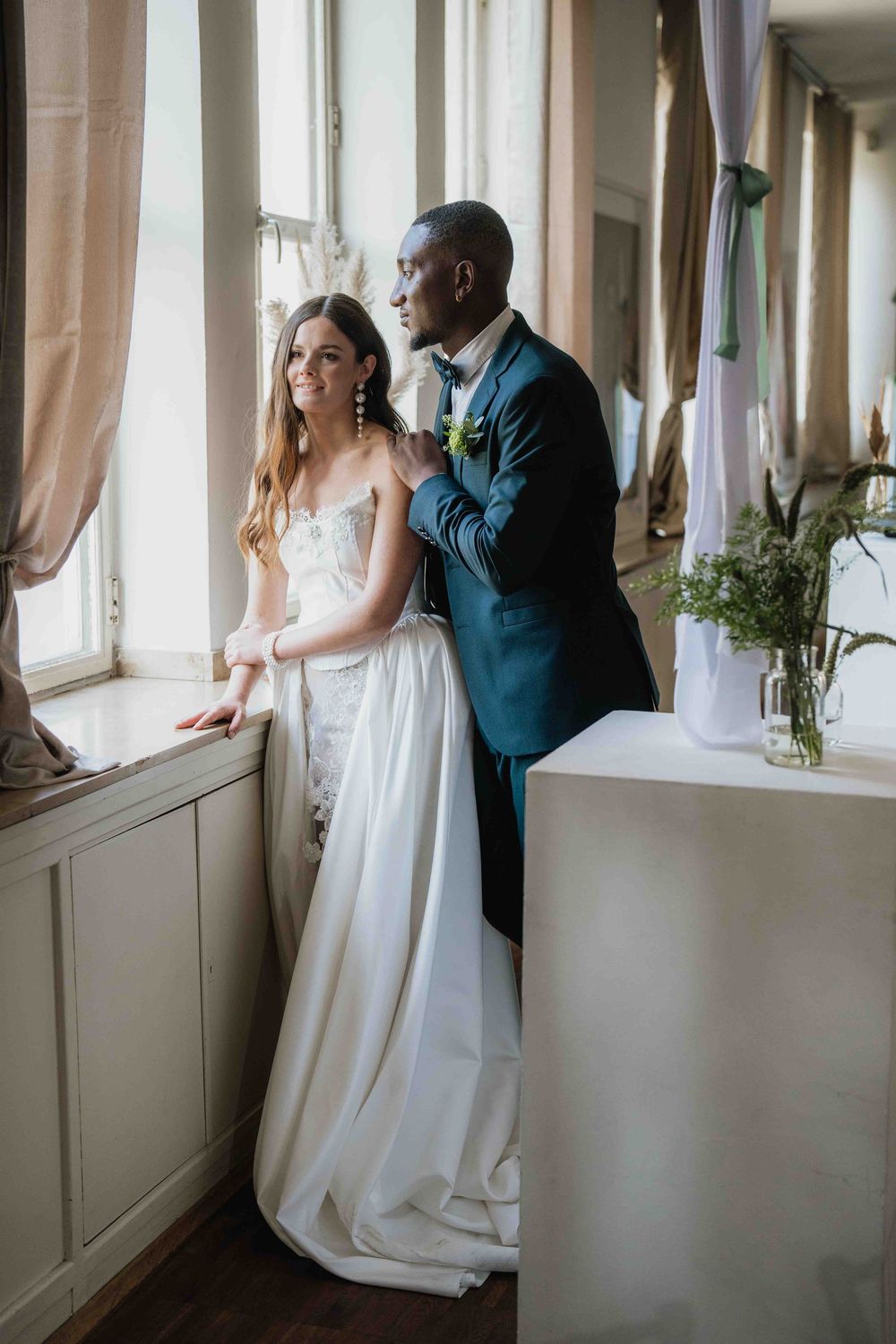A couple in wedding attire share an intimate moment by a window in an elegant indoor setting.
