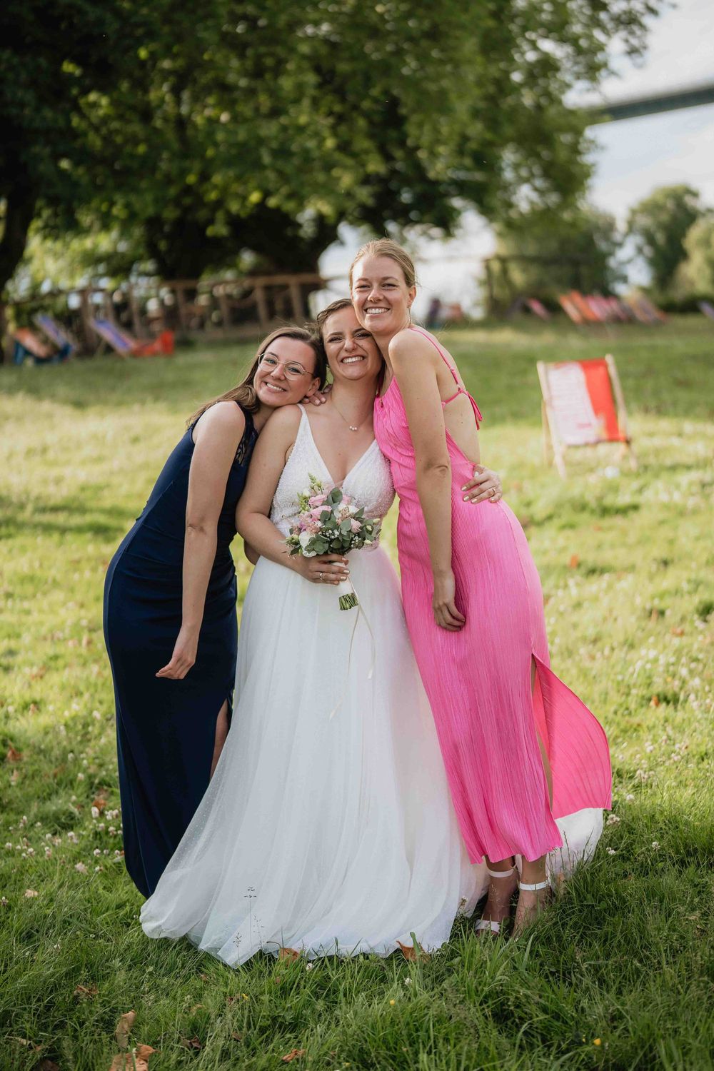 Three women in formal dresses share a celebratory moment on green grass.