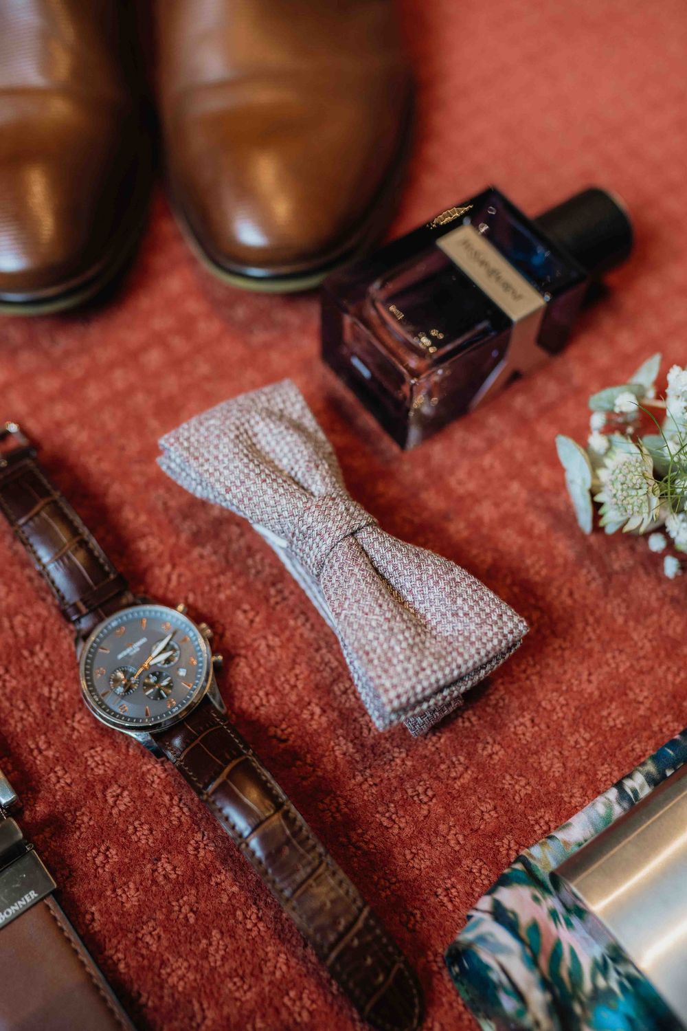 Wedding accessories including brown leather shoes, watch, bowtie and cologne on red fabric.