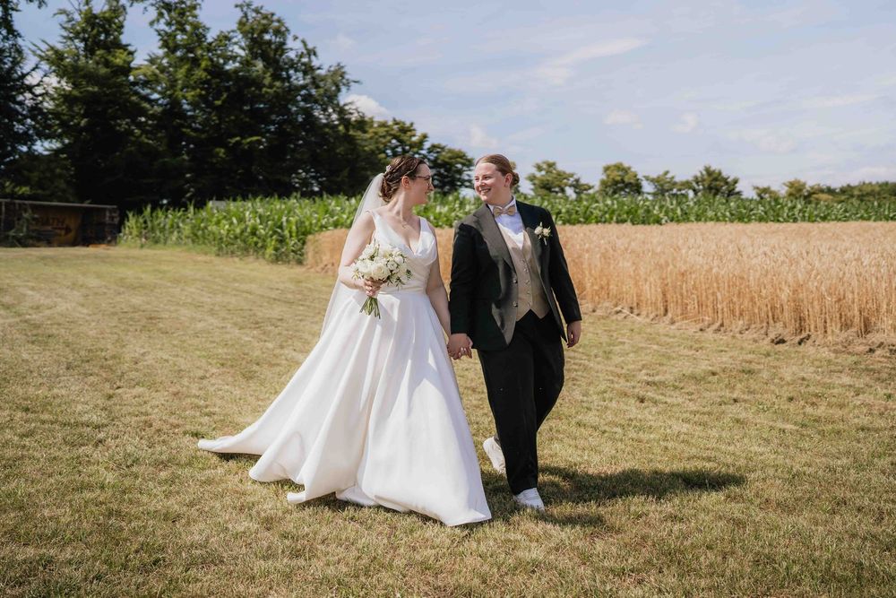 A wedding couple walking together across a sunny field with crops in the background.