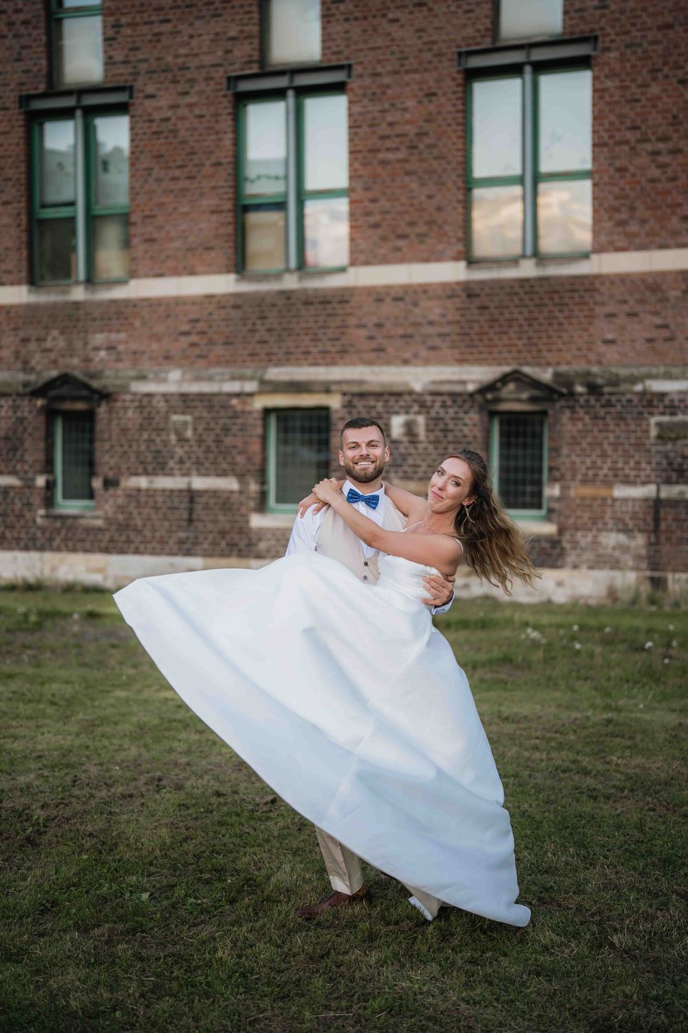 A groom lifts his bride in a flowing white dress on a grassy lawn.
