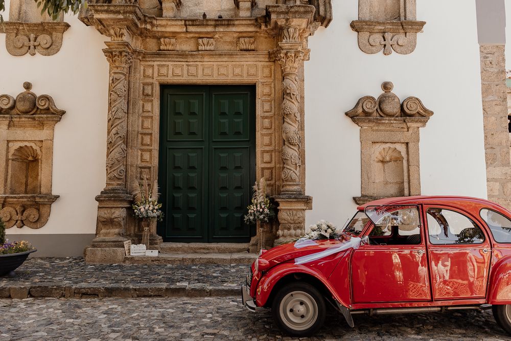 Red vintage Citroen 2CV parked on cobblestones before ornate baroque church doorway with green doors.