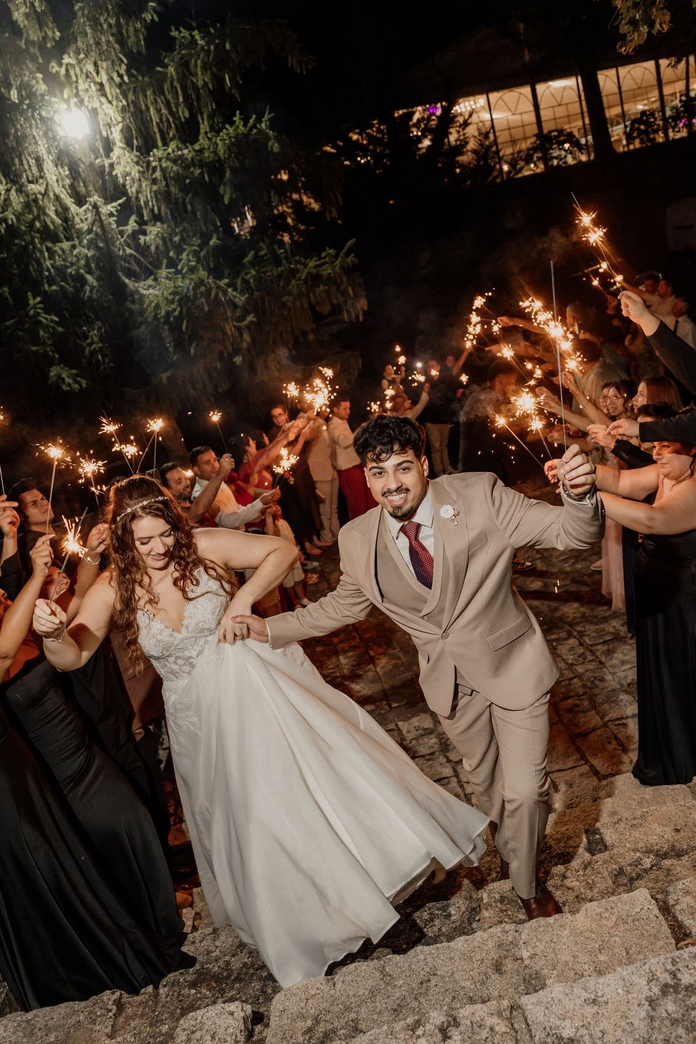 Bride and groom exit wedding reception through sparkler tunnel on stone steps at night.