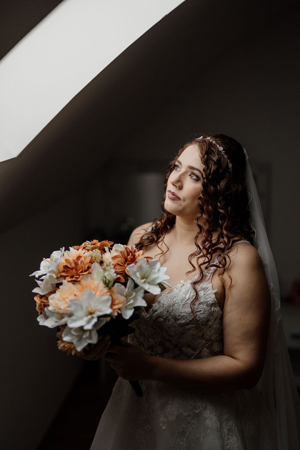 Bride in lace gown holding orange and white floral bouquet, gazing upward near a window in soft natural light.