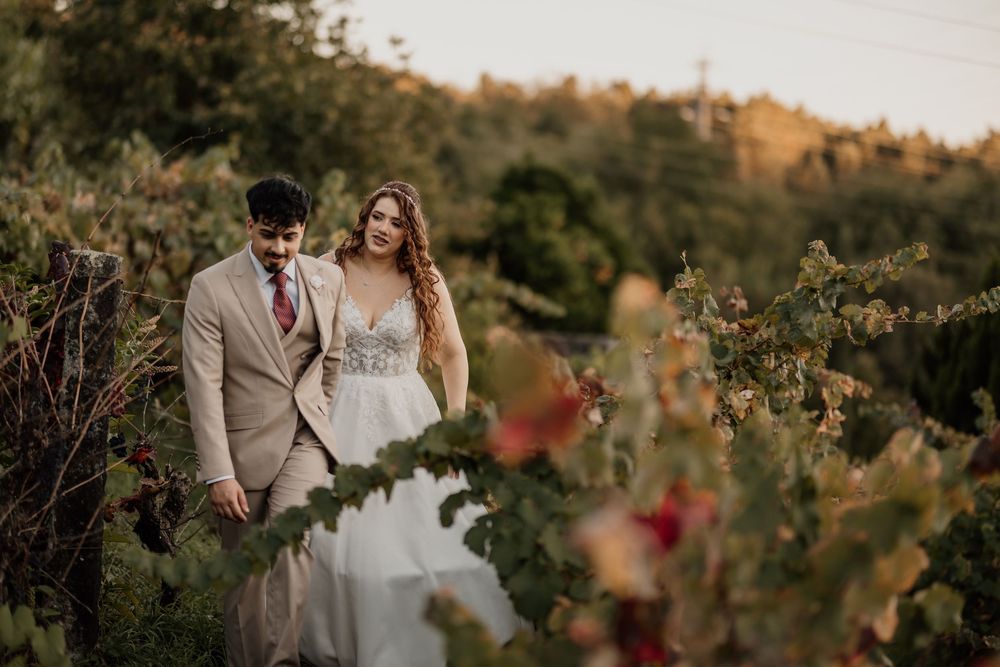 Couple walks through lush vineyard at golden hour, bride in floral gown, groom in tan suit, romantic outdoor wedding.