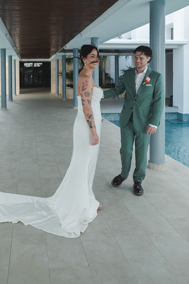 A person in a wedding dress and green-suited figure walking on gray tiles in a covered walkway space.