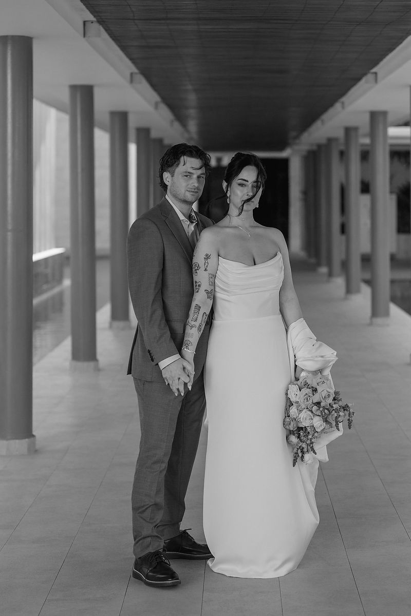 A couple poses in a modern hallway with columns, the bride wearing an elegant white dress holding a floral bouquet.