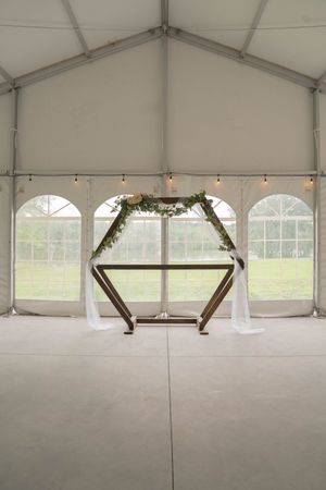 A modern geometric wedding arch with greenery stands in a white tent venue.
