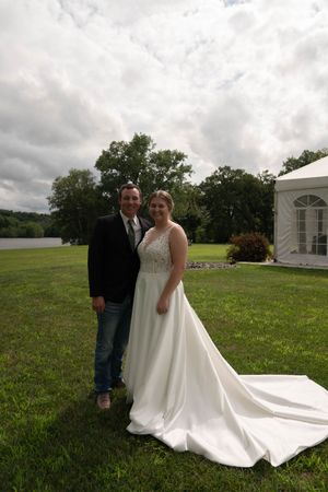 Newlyweds standing on grass with white tent and cloudy sky in background.