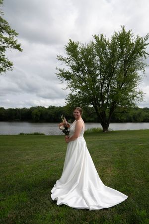 A bride in a flowing white wedding dress poses by a lake with trees and cloudy skies in the background.