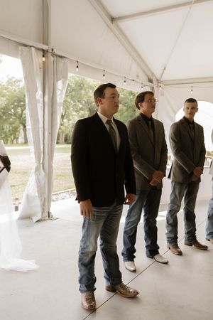 Groomsmen lined up in dark suits and jeans inside a white wedding tent.