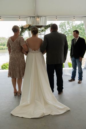A wedding party stands in a tent venue with the bride's elegant white dress train flowing behind her.