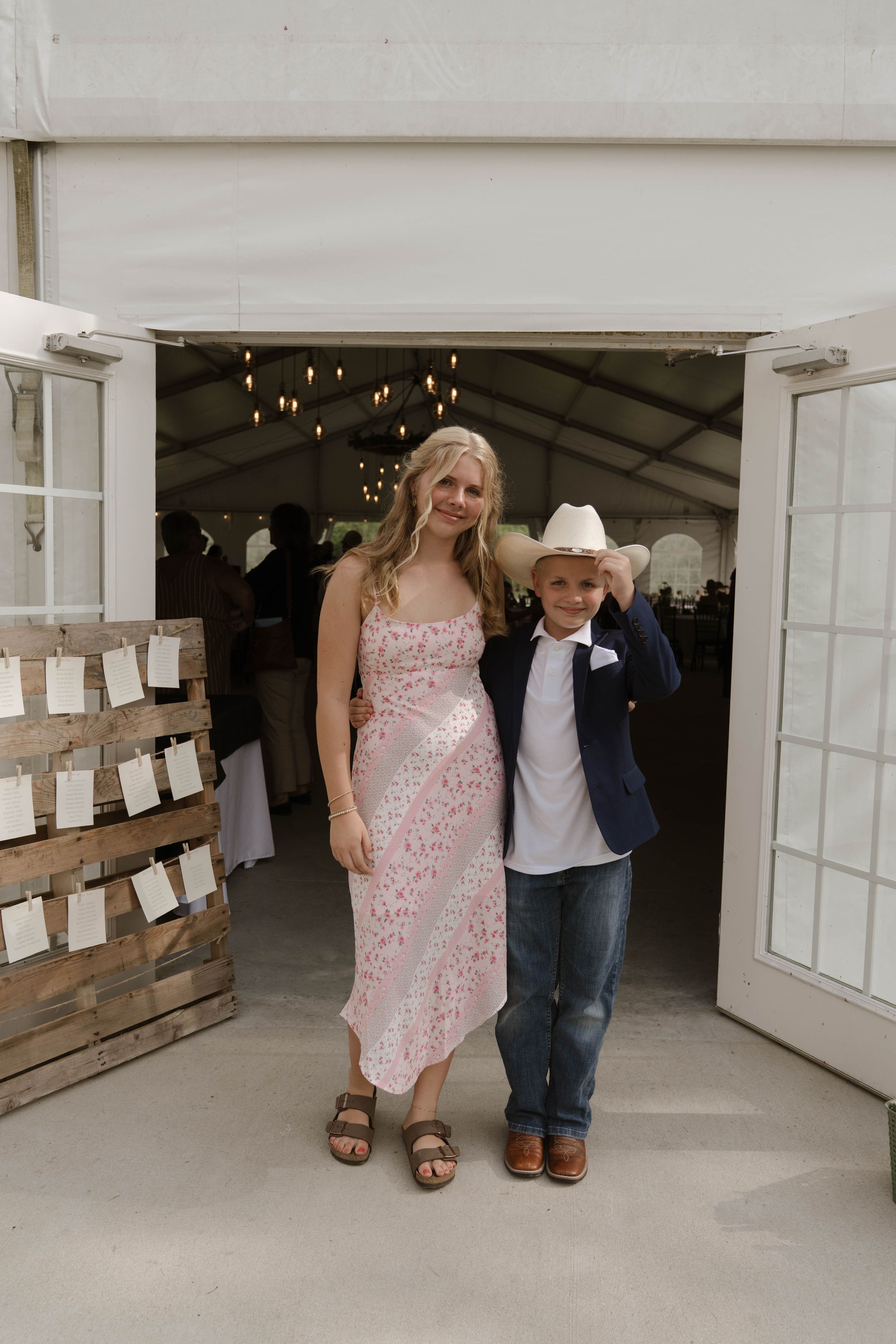Two young attendees in formal wear pose by rustic wooden doors at a wedding venue.