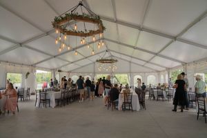 Wedding reception setup in a white tent with rustic chandeliers and dark wooden chairs.