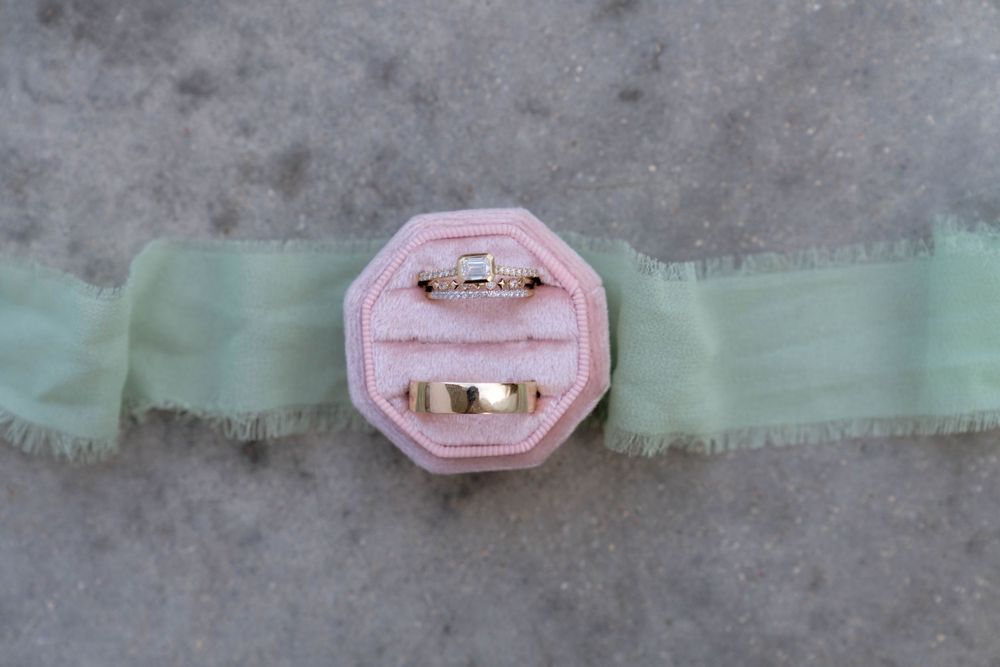 Pink velvet ring box with gold trim displayed on mint green ribbon against concrete background.