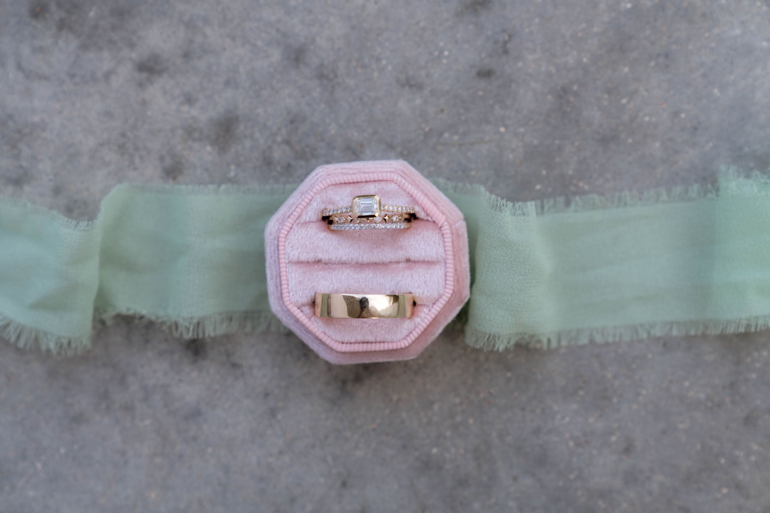 Pink velvet ring box with gold trim displayed on mint green ribbon against concrete background.
