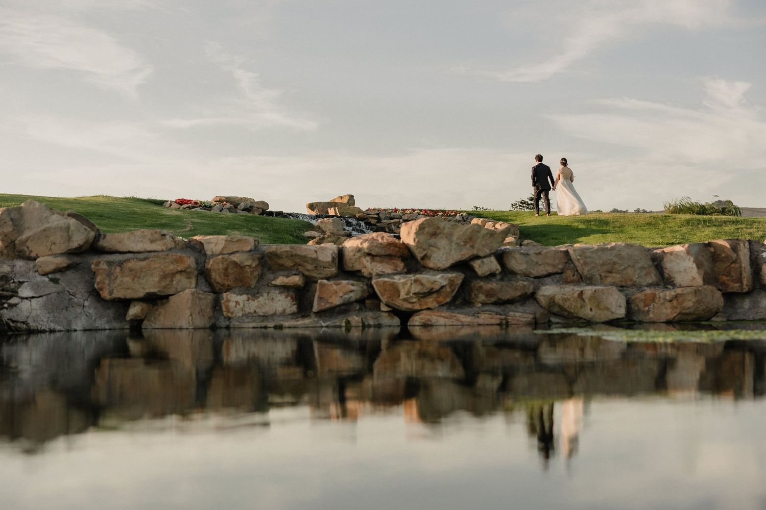 A silhouetted figure stands on rocky outcrops by a serene lake during sunset creating a peaceful panoramic scene.
