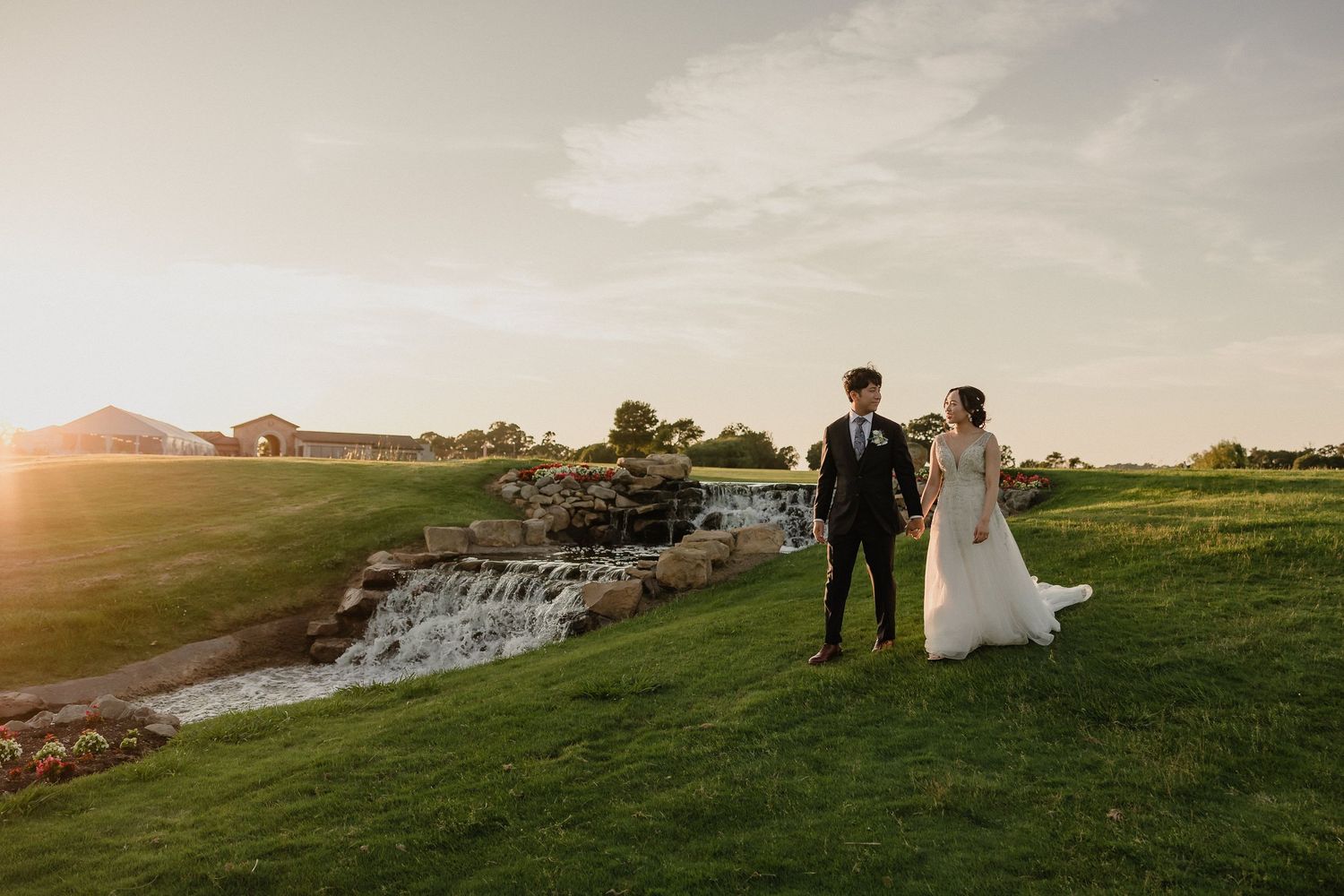Two people in wedding attire stand on a grassy hilltop overlooking a scenic waterfall at sunset.