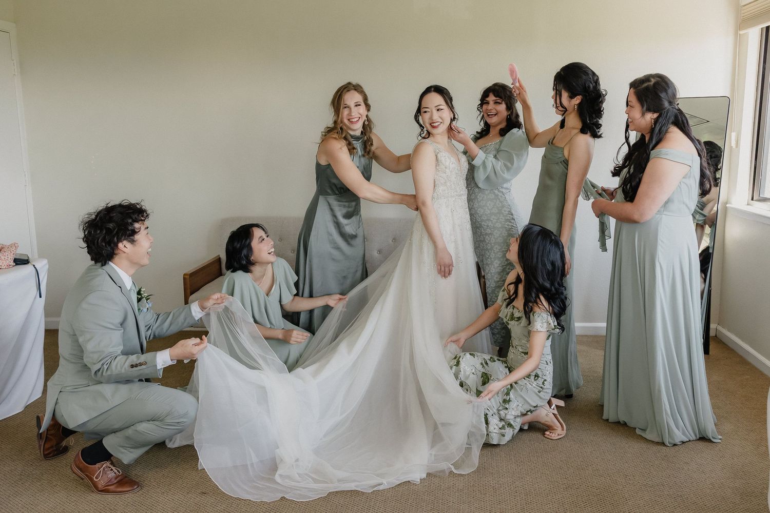 Bridesmaids in sage green dresses help adjust the flowing white wedding gown during wedding preparations.