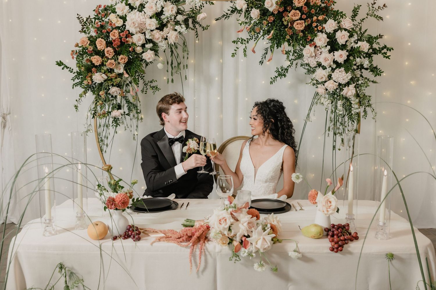 Newlyweds share a toast at an elegant reception table decorated with lush floral arrangements and romantic archways.