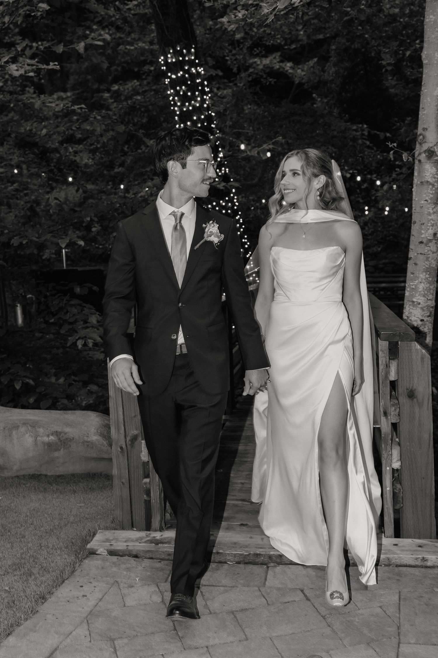 Black and white photo of newlyweds walking together outdoors with string lights and trees in background.