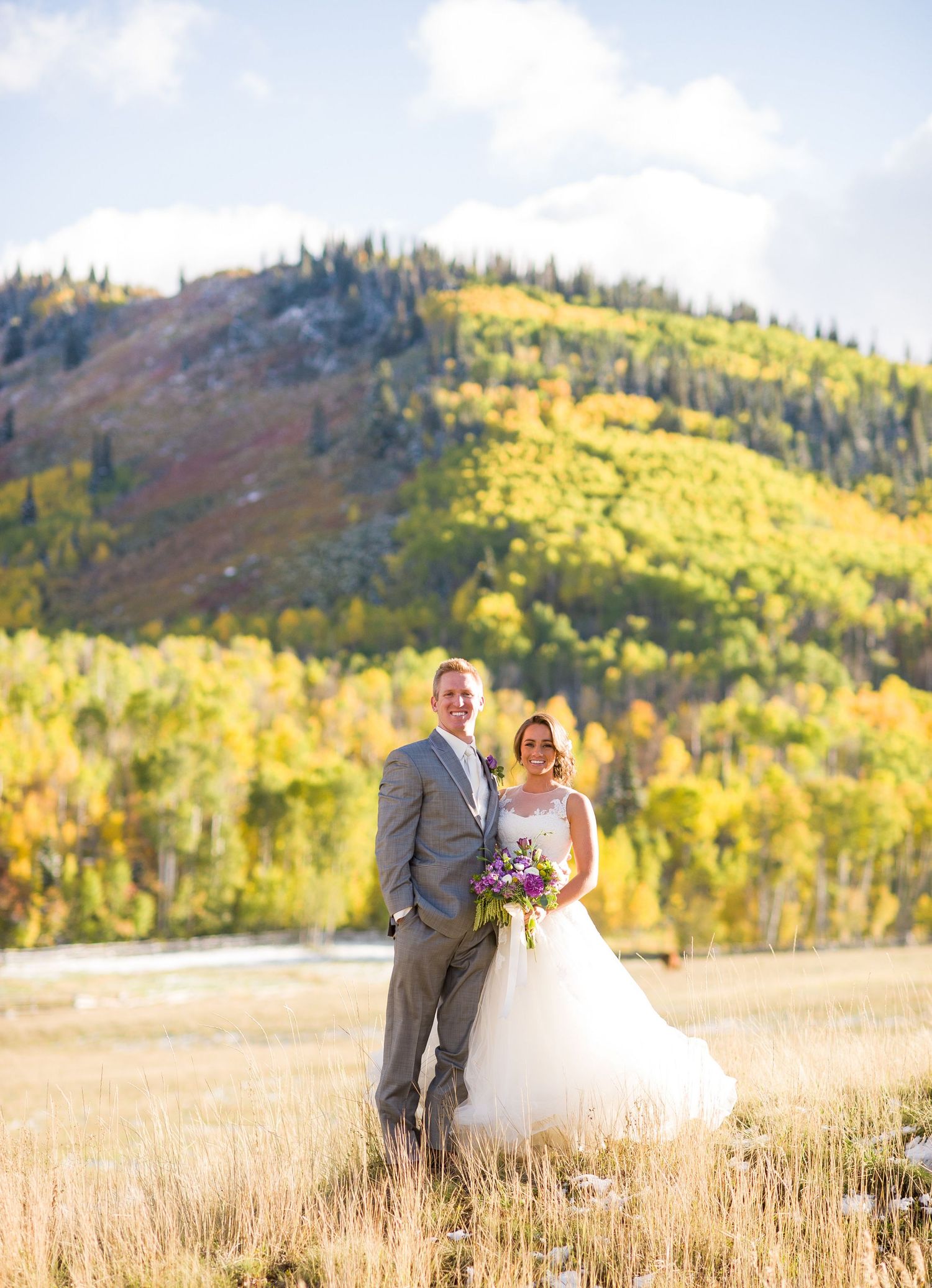 Newlyweds standing in mountain meadow with autumn foliage and sunlit hillside behind them.