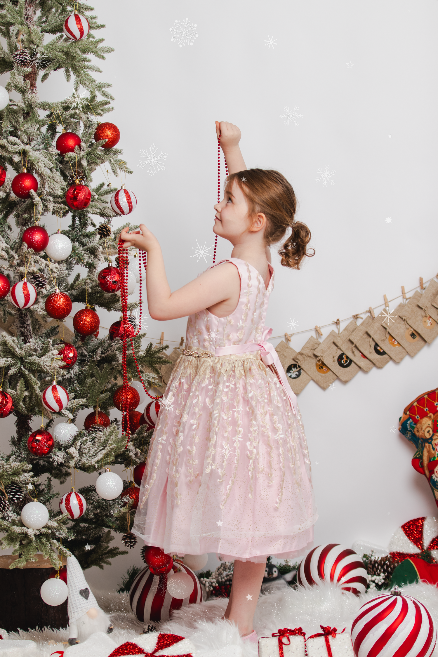 A child in a pink party dress decorates a Christmas tree with red and white ornaments.