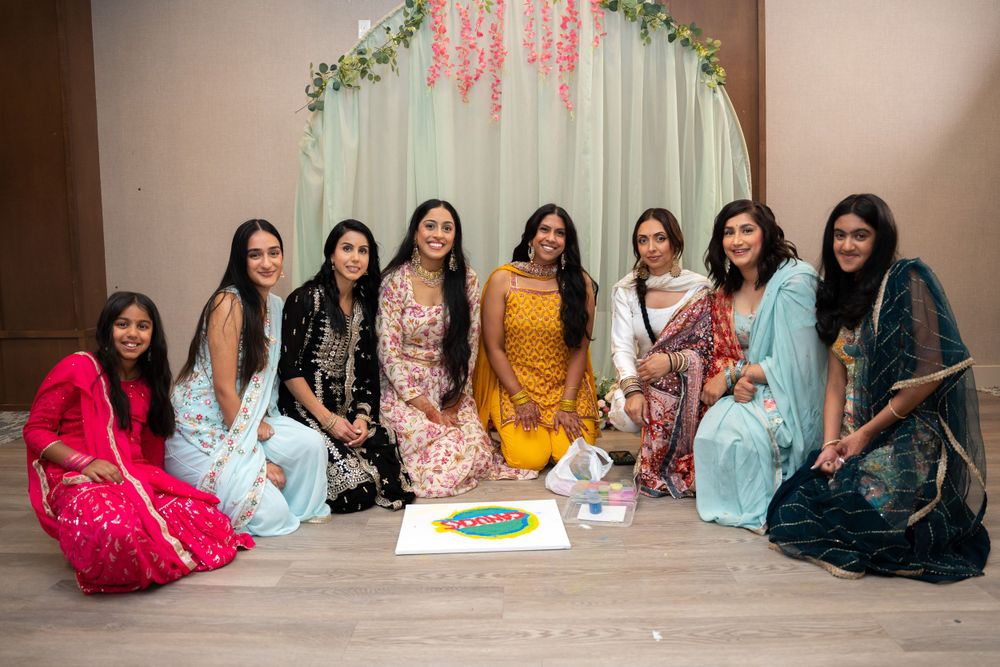A festive gathering of friends in traditional Indian attire seated on the floor around decorative items.