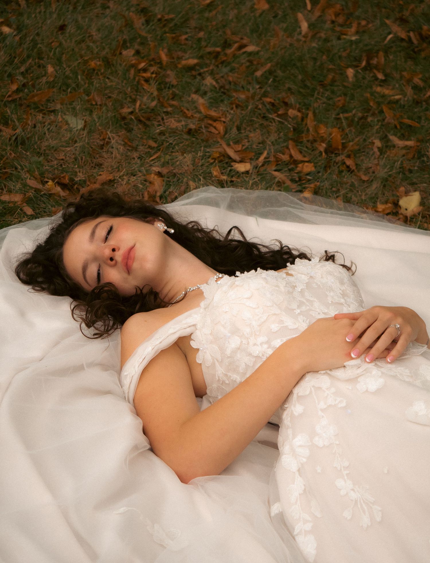 A bride in lace dress lies peacefully on white fabric surrounded by fallen leaves.