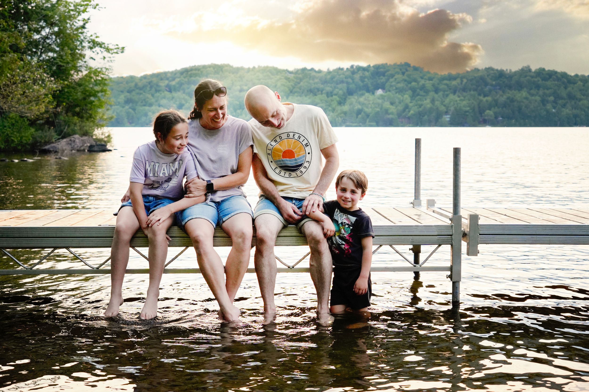 Seance photo de famille avec de jeunes enfants assis sur un quai les pieds dans l'eau au golden hour à Orford, Quebec