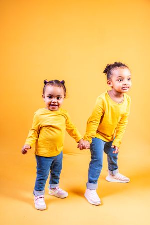Two toddlers in matching yellow shirts and jeans hold hands while posing against orange background.