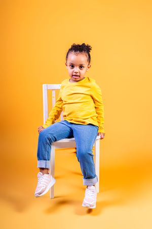 Child in bright yellow shirt and jeans sits on white chair against orange background.