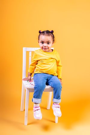 Toddler in yellow top and jeans sits casually on white chair against orange backdrop.