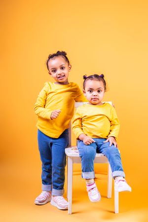 Two children in matching yellow shirts pose together with white chair against orange background.