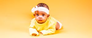 Baby in yellow outfit wearing white headband lying on orange background.