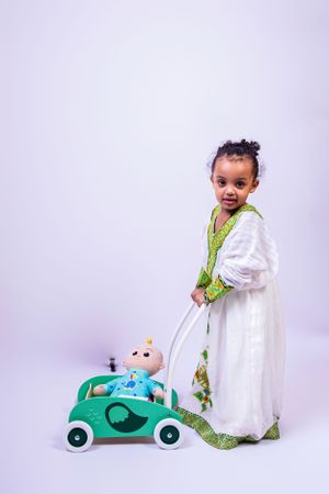 Small child in traditional white dress pushing a green toy car with a doll.