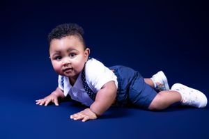 A baby in blue overalls crawls on a dark blue studio backdrop.