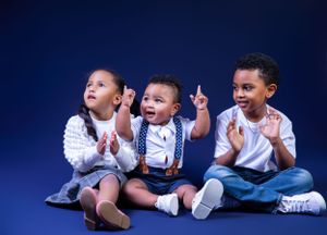 Three children in matching white shirts and suspenders gesture excitedly against a blue background.