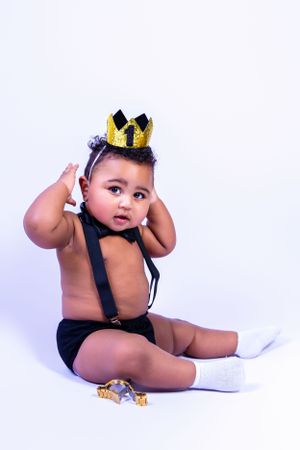 Baby wearing black suspenders and gold crown poses while sitting in white studio setting with toys.