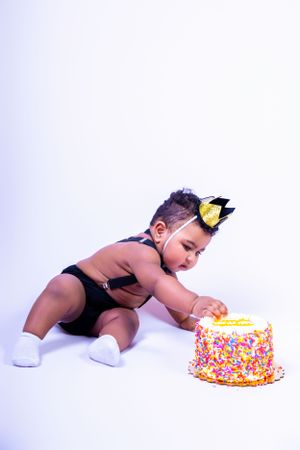 Baby in black outfit and crown reaches for colorful birthday cake against white background.
