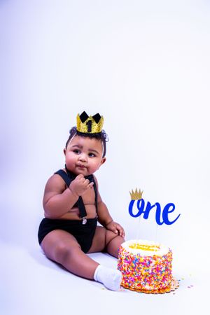 Baby in black outfit and crown sits next to first birthday cake with blue 'One' decoration.